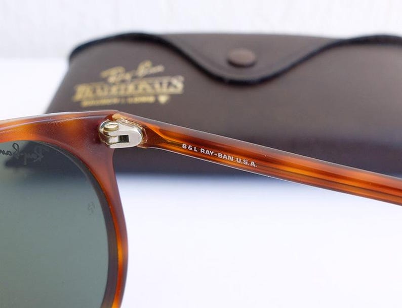 Puede incluir: Primer plano de gafas de sol Ray-Ban con montura carey y lentes verdes. La patilla derecha est&aacute; marcada con "B&L RAY-BAN U.S.A.". En el fondo, un estuche de cuero marr&oacute;n con letras doradas.