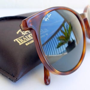 Puede incluir: Gafas de sol carey con lentes oscuras, que reflejan un cielo azul y una palmera. El logotipo de Ray-Ban es visible en la montura. Un estuche de cuero marr&oacute;n oscuro con letras doradas est&aacute; en el fondo.