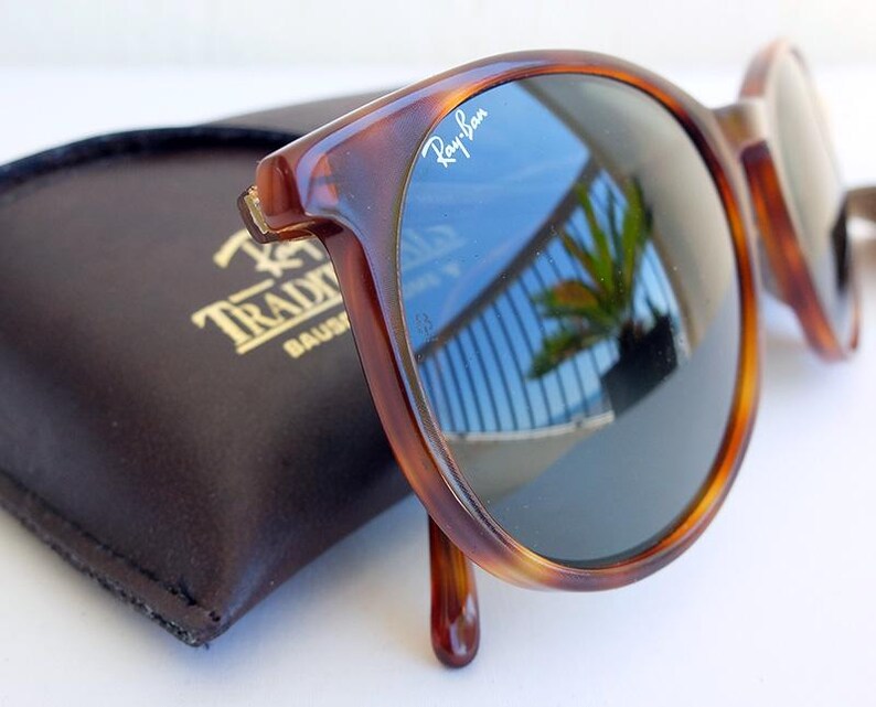Puede incluir: Gafas de sol carey con lentes oscuras, que reflejan un cielo azul y una palmera. El logotipo de Ray-Ban es visible en la montura. Un estuche de cuero marr&oacute;n oscuro con letras doradas est&aacute; en el fondo.