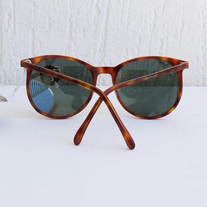 Puede incluir: Gafas de sol redondas con montura carey y lentes verde oscuro. Las patillas de las gafas est&aacute;n extendidas, mostrando la parte trasera de la montura. Las gafas est&aacute;n sobre una superficie blanca.