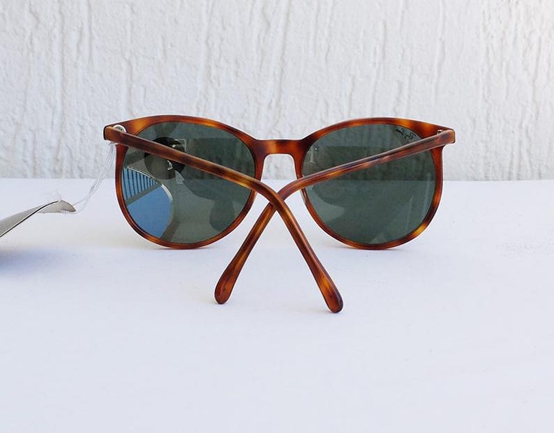 Puede incluir: Gafas de sol redondas con montura carey y lentes verde oscuro. Las patillas de las gafas est&aacute;n extendidas, mostrando la parte trasera de la montura. Las gafas est&aacute;n sobre una superficie blanca.