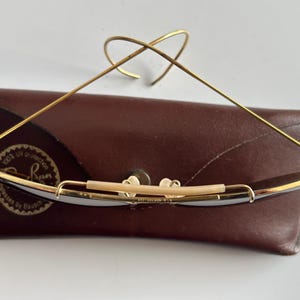 K&ouml;nnte beinhalten: Goldgerahmte Sonnenbrille mit dunklen Gl&auml;sern liegt auf einem braunen Lederetui. Die Brille hat ein markantes Drahtdesign und einen gebogenen oberen B&uuml;gel. Das Etui hat ein Logo und Text.
