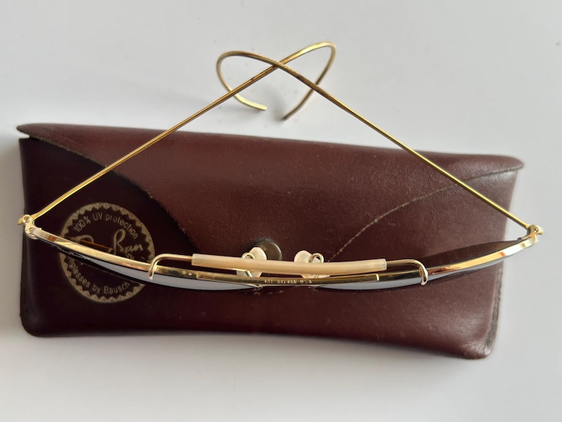 K&ouml;nnte beinhalten: Goldgerahmte Sonnenbrille mit dunklen Gl&auml;sern liegt auf einem braunen Lederetui. Die Brille hat ein markantes Drahtdesign und einen gebogenen oberen B&uuml;gel. Das Etui hat ein Logo und Text.