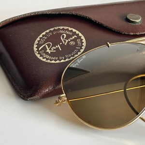 K&ouml;nnte beinhalten: Goldgerahmte Ray-Ban Sonnenbrille mit braunen Gl&auml;sern, neben einem braunen Lederetui. Das Etui zeigt das Ray-Ban-Logo und den Text, der 100 % UV-Schutz anzeigt. Die Sonnenbrille hat ein klassisches Aviator-Design.