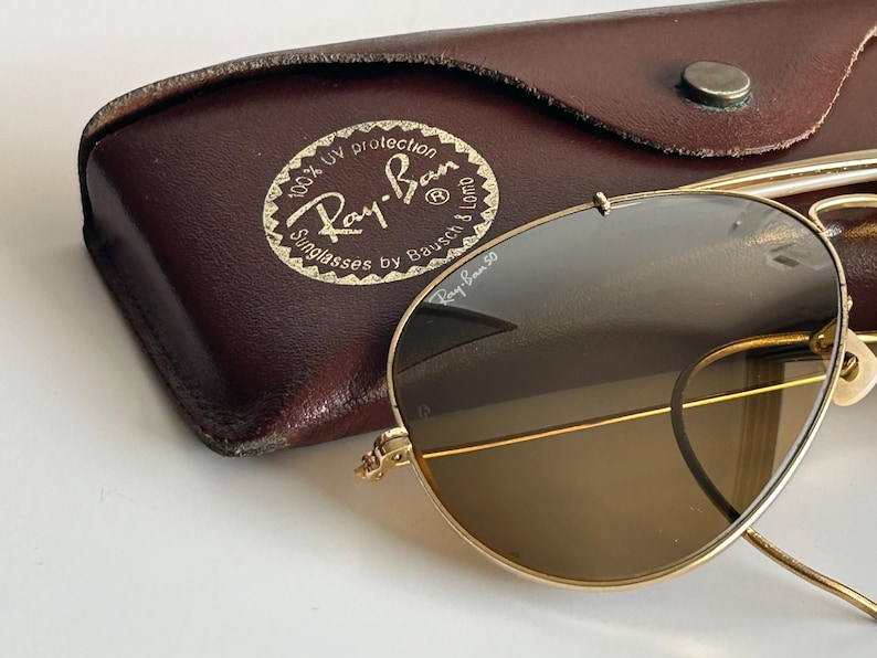 K&ouml;nnte beinhalten: Goldgerahmte Ray-Ban Sonnenbrille mit braunen Gl&auml;sern, neben einem braunen Lederetui. Das Etui zeigt das Ray-Ban-Logo und den Text, der 100 % UV-Schutz anzeigt. Die Sonnenbrille hat ein klassisches Aviator-Design.