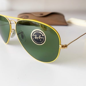 Puede incluir: Gafas de sol con montura amarilla y lentes verdes. Las gafas tienen el logotipo de Ray-Ban y el texto "G-15 ULTRA VIOLET PROTECTION" en la lente derecha.
