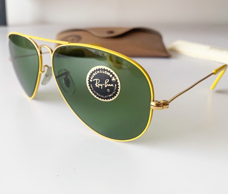 Puede incluir: Gafas de sol con montura amarilla y lentes verdes. Las gafas tienen el logotipo de Ray-Ban y el texto "G-15 ULTRA VIOLET PROTECTION" en la lente derecha.