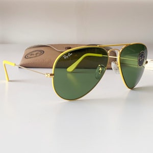 Puede incluir: Gafas de sol aviador amarillas con lentes verdes. Las gafas tienen un marco dorado y el logotipo de Ray-Ban es visible en la lente derecha.