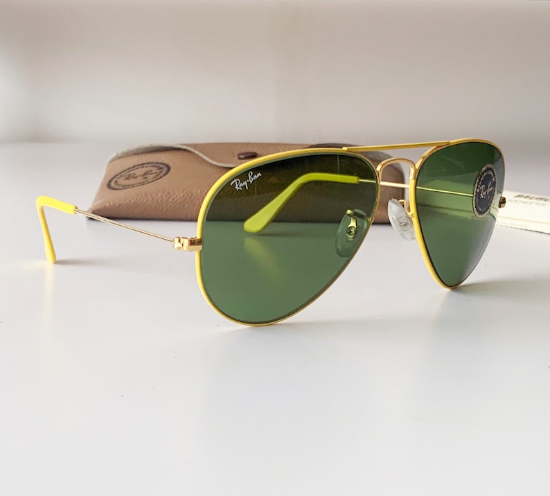 Puede incluir: Gafas de sol aviador amarillas con lentes verdes. Las gafas tienen un marco dorado y el logotipo de Ray-Ban es visible en la lente derecha.