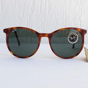 Puede incluir: Gafas de sol redondas con montura carey y lentes verde oscuro. El logotipo de Ray-Ban es visible en la parte superior izquierda de la montura y en la lente. Una peque&ntilde;a etiqueta est&aacute; adherida a la montura.