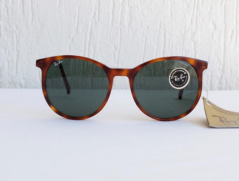 Puede incluir: Gafas de sol redondas con montura carey y lentes verde oscuro. El logotipo de Ray-Ban es visible en la parte superior izquierda de la montura y en la lente. Una peque&ntilde;a etiqueta est&aacute; adherida a la montura.