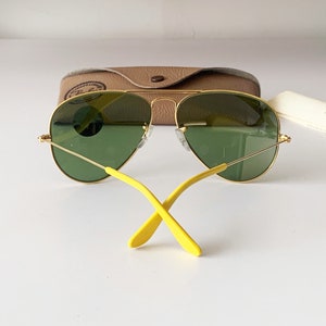 Puede incluir: Gafas de sol aviador doradas con lentes verdes y brazos amarillos. Las gafas de sol est&aacute;n apoyadas en una caja marr&oacute;n.