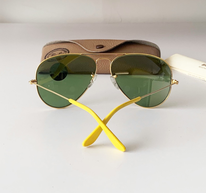 Puede incluir: Gafas de sol aviador doradas con lentes verdes y brazos amarillos. Las gafas de sol est&aacute;n apoyadas en una caja marr&oacute;n.