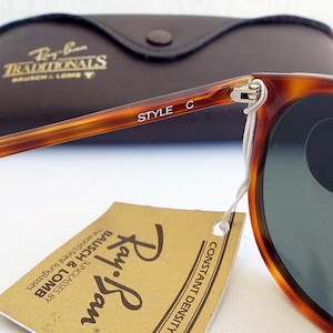 Puede incluir: Primer plano de gafas de sol Ray-Ban con montura carey y lentes verdes. Las palabras "STYLE C" est&aacute;n impresas en la patilla. Tambi&eacute;n se ve un estuche de cuero marr&oacute;n y una etiqueta con el logotipo de Ray-Ban.