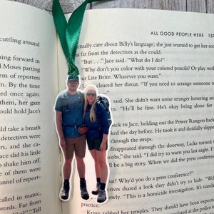 May include: A green and white bookmark with a photo of a couple standing together. The bookmark is shaped like a rectangle with rounded corners. The photo is of a couple standing in front of a mountain range. The bookmark is attached to a green ribbon.