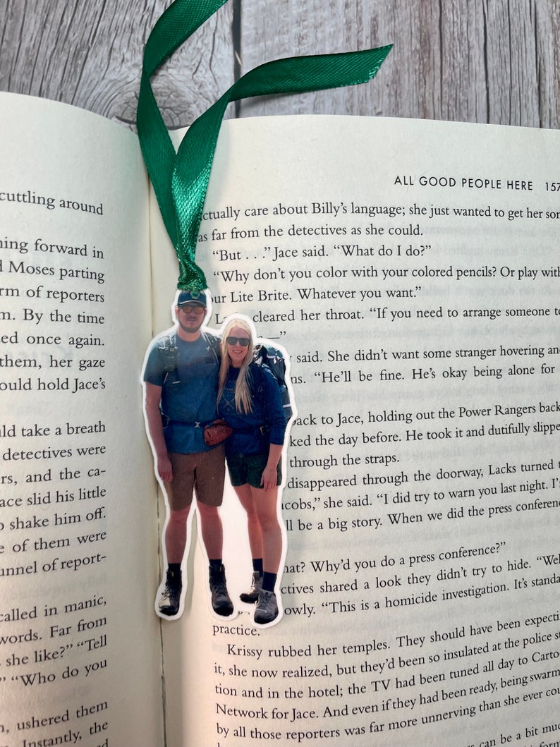 May include: A green and white bookmark with a photo of a couple standing together. The bookmark is shaped like a rectangle with rounded corners. The photo is of a couple standing in front of a mountain range. The bookmark is attached to a green ribbon.
