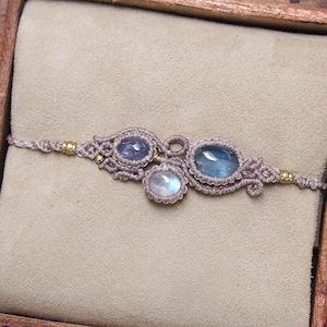 May include: A macrame bracelet featuring three oval-shaped gemstones in shades of blue and iridescent white, set within intricate beige knotted cord. The design includes small gold-toned metal beads integrated into the cord on both sides.
