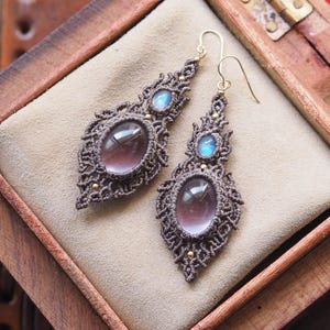 May include: A pair of macrame earrings featuring brown cord woven into intricate patterns. Each earring holds a large oval purple gemstone and a smaller oval blue gemstone. The earrings are finished with gold-toned metal hooks and small gold beads.