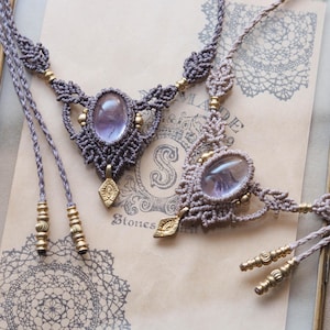 May include: Two macrame necklaces with oval, light purple stones. The necklaces are grey with gold accents and leaf-shaped pendants. The background features a framed vintage paper with decorative patterns and the word "Stones".