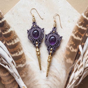 May include: Handmade macrame earrings with an oval amethyst stone. The earrings have a dark purple macrame design, gold-coloured accents, and a gold-coloured hook. They are displayed on a light-coloured stone surface, with feathers in the background.