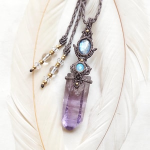 Op de afbeelding: Een macramé ketting met een grote, verticale, doorschijnende paarse amethist kristalpunt. De hanger is versierd met een kleine, ronde, iriserende blauwe steen en een ovale, gefacetteerde blauwe steen, gezet in donkerbruin geknoopt koord.