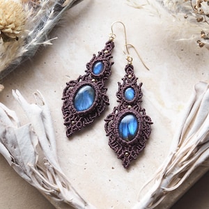 Puede incluir: Un par de pendientes colgantes con cordón de macramé marrón oscuro tejido en patrones intrincados alrededor de piedras preciosas de labradorita ovaladas. Cada pendiente muestra dos piedras azules iridiscentes y un gancho de metal dorado.