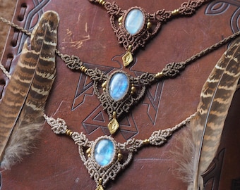 Puede incluir: Tres collares de macramé con piedras preciosas ovaladas de color azul claro y detalles dorados se exhiben sobre un libro encuadernado en cuero marrón. Los collares están hechos de cordones marrones y beige. Hay plumas a ambos lados de los collares.