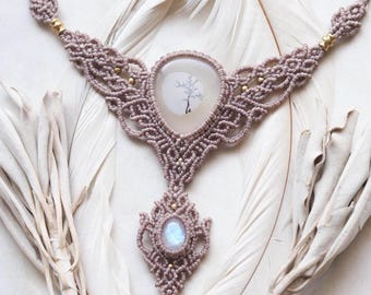 May include: A delicate macrame necklace with a light brown cord and gold beads. The necklace features a large, oval-shaped pendant with a clear stone and a tree silhouette inside. A smaller, teardrop-shaped pendant with a moonstone hangs below.