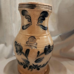 May include: A tan stoneware pitcher with a salt-glaze finish and cobalt blue hand-painted floral motifs. The front features a circular stamp reading HANDMADE ROWE POTTERY WORKS CAMBRIDGE WIS LIMITED PRODUCTION SERIES.