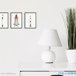 May include: Set of three black and white illustrations of rockets and space shuttles. The illustrations are framed and displayed on a white wall. The frames are 5x7 inches.