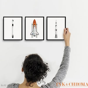 May include: Set of three black framed prints featuring illustrations of different types of rockets. The prints are hanging on a white wall. The prints are 8x10 inches.