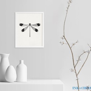 May include: A black and white illustration of a dragonfly in a white frame, with two white vases and a branch with white flowers on a white surface.