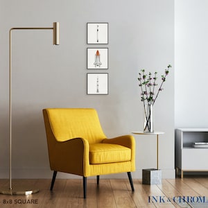 May include: A set of three 8x8 inch square prints featuring minimalist illustrations of a rocket, a space shuttle, and a rocket with a trail of smoke. The prints are displayed on a white wall in a living room setting with a yellow armchair, a floor lamp, a vase of flowers, and a TV stand.