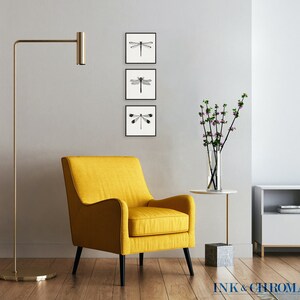 May include: A yellow armchair with black legs sits in a living room with a floor lamp, a small table with a vase of flowers, and a television on a white cabinet. Three black and white framed prints of dragonflies hang on the wall.