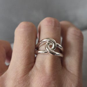 May include: A silver ring with a knot design. The ring has a smooth, polished surface and a unique, intertwined knot shape. The ring is shown on a finger, highlighting its elegant and minimalist style.