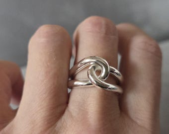 May include: A silver-colored ring with a knot design. The ring features a smooth, polished surface and a unique, intertwined knot shape. The ring is shown on a finger, highlighting its elegant and minimalist style.