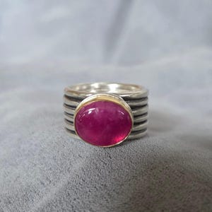 May include: A wide sterling silver ring featuring a prominent oval-shaped, deep pink cabochon gemstone set in a thin gold-toned bezel. The band is constructed from multiple stacked, horizontal silver wires with darkened, oxidized recessed grooves.