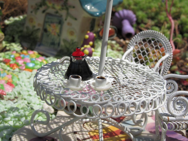 Peut inclure: Une table en m&eacute;tal blanc avec une chaise en m&eacute;tal blanc. Il y a une cafeti&egrave;re miniature et deux tasses de caf&eacute; sur la table. La table et la chaise sont d&eacute;cor&eacute;es d'un motif d&eacute;licat et complexe.