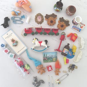May include: A collection of miniature toys and trinkets, including a red and green wooden train, a white rocking horse, and a framed portrait. Other items include a baby bottle, a pair of baby shoes, and a Hello Kitty pin.