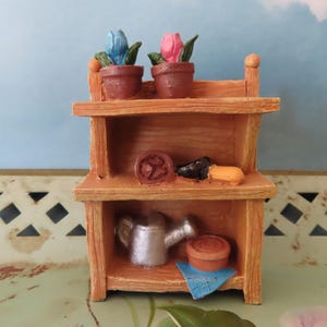 May include: A miniature wooden shelf unit with two levels. The top shelf holds two potted flowers, one blue and one pink. The middle shelf has a small brown pot, a black shoe, and a yellow item. The bottom shelf has a silver watering can, a small pot, and a blue cloth.