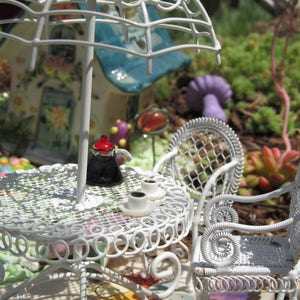 Peut inclure: Un ensemble de patio en m&eacute;tal blanc avec une table ronde et deux chaises. La table a un motif de dentelle blanche et une cafeti&egrave;re noire dessus. Il y a deux tasses et soucoupes en c&eacute;ramique blanche sur la table. Les chaises ont un motif de treillis blanc.