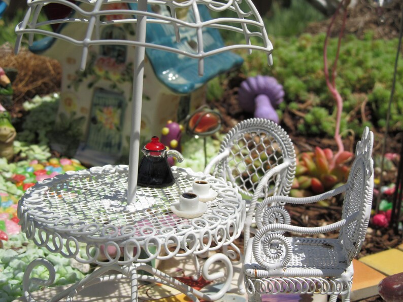 Peut inclure: Un ensemble de patio en m&eacute;tal blanc avec une table ronde et deux chaises. La table a un motif de dentelle blanche et une cafeti&egrave;re noire dessus. Il y a deux tasses et soucoupes en c&eacute;ramique blanche sur la table. Les chaises ont un motif de treillis blanc.