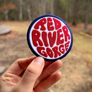 Puede incluir: Un parche circular con las palabras "RED RIVER GORGE" en letras rojas redondeadas sobre un fondo blanco. El parche tiene un borde azul, lo que sugiere que es un artículo bordado.