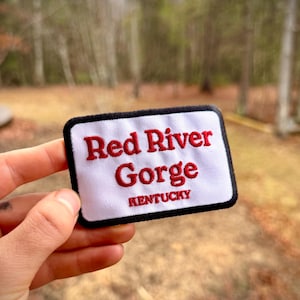 May include: A rectangular white patch with black trim features the words "Red River Gorge" in red embroidered letters, with "Kentucky" below. The patch is held up against a blurred outdoor background.