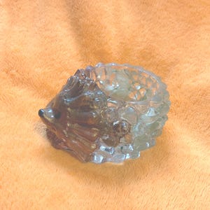May include: A clear glass hedgehog-shaped container. The container has a detailed design, with a textured body and a smooth, rounded head. The glass has a slight tint of green and brown, and the container is on an orange surface.