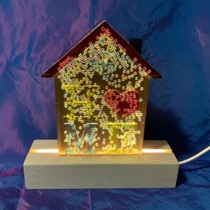 May include: A house-shaped night light with a clear base and a wooden stand. The house is filled with iridescent glitter and the word "HOME" is spelled out in the centre with a red heart.