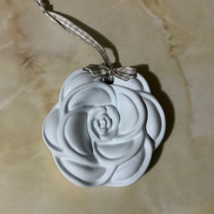 May include: A white ceramic rose ornament with a beige ribbon attached. The rose is detailed with a swirl pattern and has a hole in the centre.