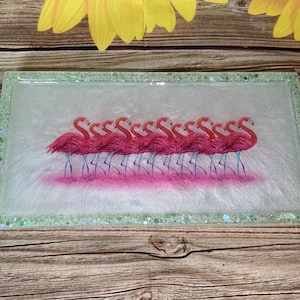May include: Rectangular resin tray with a flamingo design. The tray features a row of pink flamingos with blue legs and a pink base. The edges are outlined with glitter. The background is white with a pink hue.