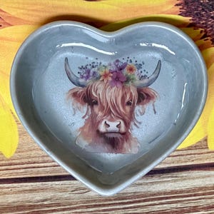 May include: A heart-shaped trinket dish in a pale grey colour. The dish has a detailed illustration of a Highland cow wearing a floral crown. The cow is brown and white with curved horns. The dish is likely made of ceramic.