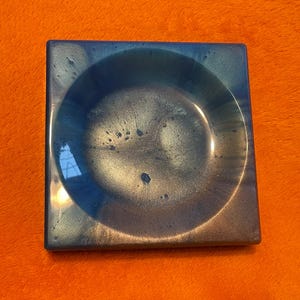 May include: A square, blue and gold resin tray with a rounded, slightly raised centre. The tray has a speckled, iridescent finish.